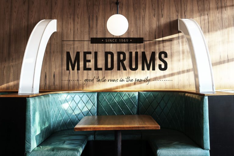 Meldrums logo on a wood-paneled wall above teal booth seating, reading ‘MELDRUMS’ and ‘good taste runs in the family’ (brand identity services provided by Rebecca Snyder, A LA MODE designs. Rebecca is an award-winning graphic designer).