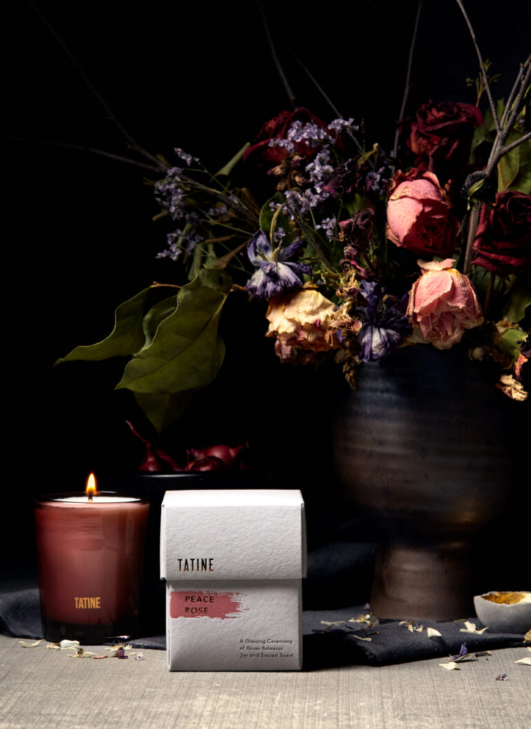 Lit TATINE candle beside the ‘Peace Rose’ packaging box on a styled tabletop with dried roses and florals in the background (package design by Rebecca Snyder, A LA MODE designs).
