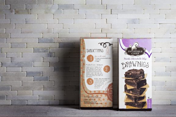 Two The Invisible Chef brownie mix boxes photographed against a light brick wall, showing the front label and back directions panel. Packaging design by Rebecca Snyder of A LA MODE designs.