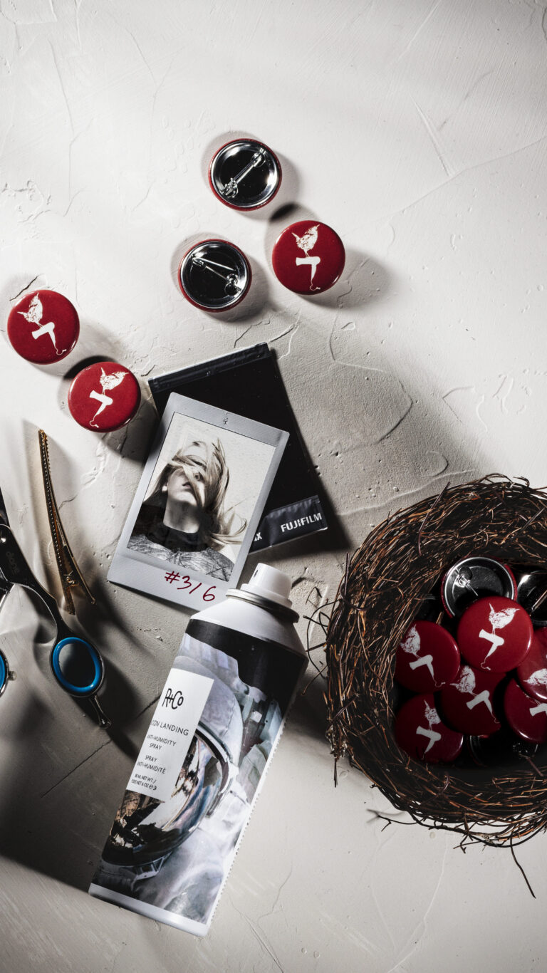 Flat lay of Red Wren Hair pinback buttons scattered on a light surface with scissors, a Fujifilm instant photo, an aerosol can, and a twig nest holding more buttons.
