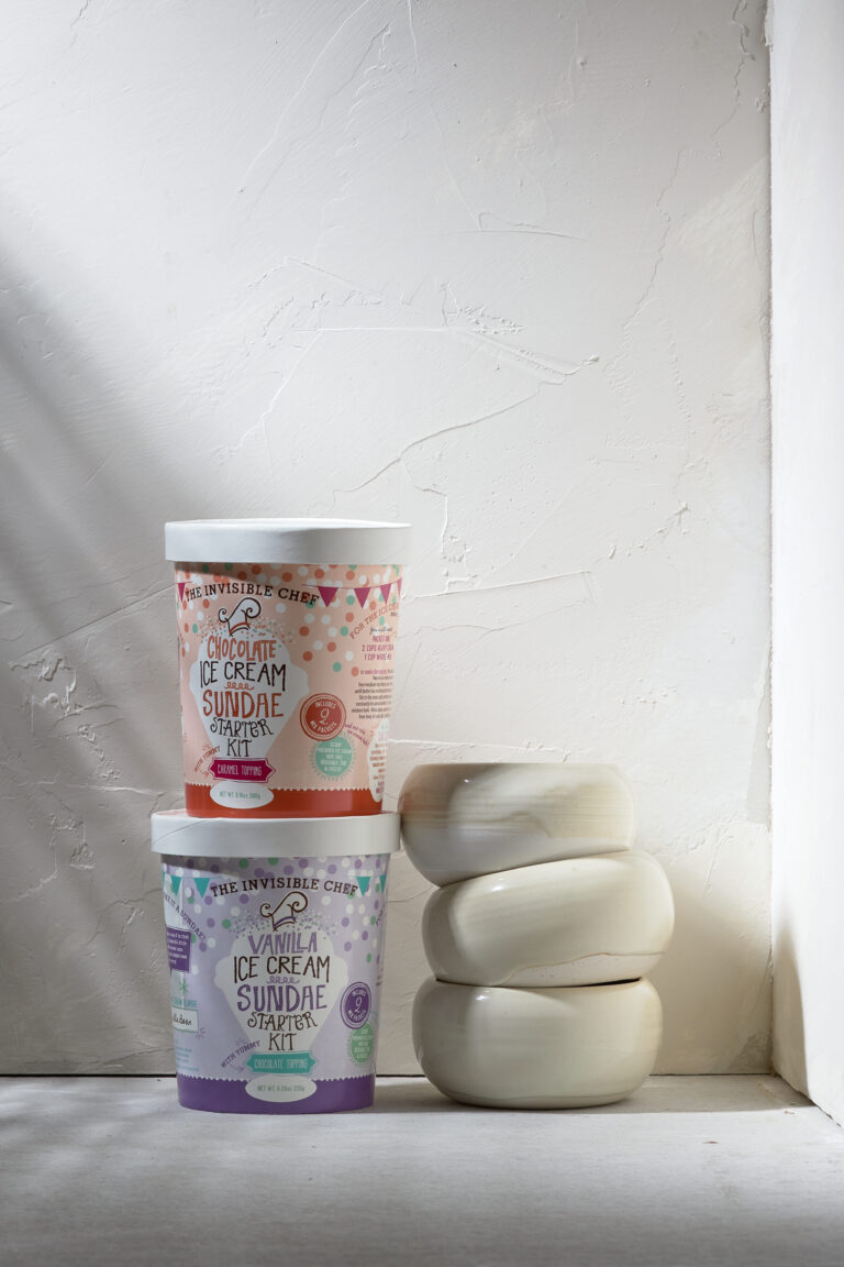 Two The Invisible Chef ice cream sundae starter kit containers (chocolate stacked on vanilla) beside stacked ceramic bowls against a textured white wall. Package design by Rebecca Snyder of A LA MODE designs, an award winning design studio, located in Canton, Ohio.