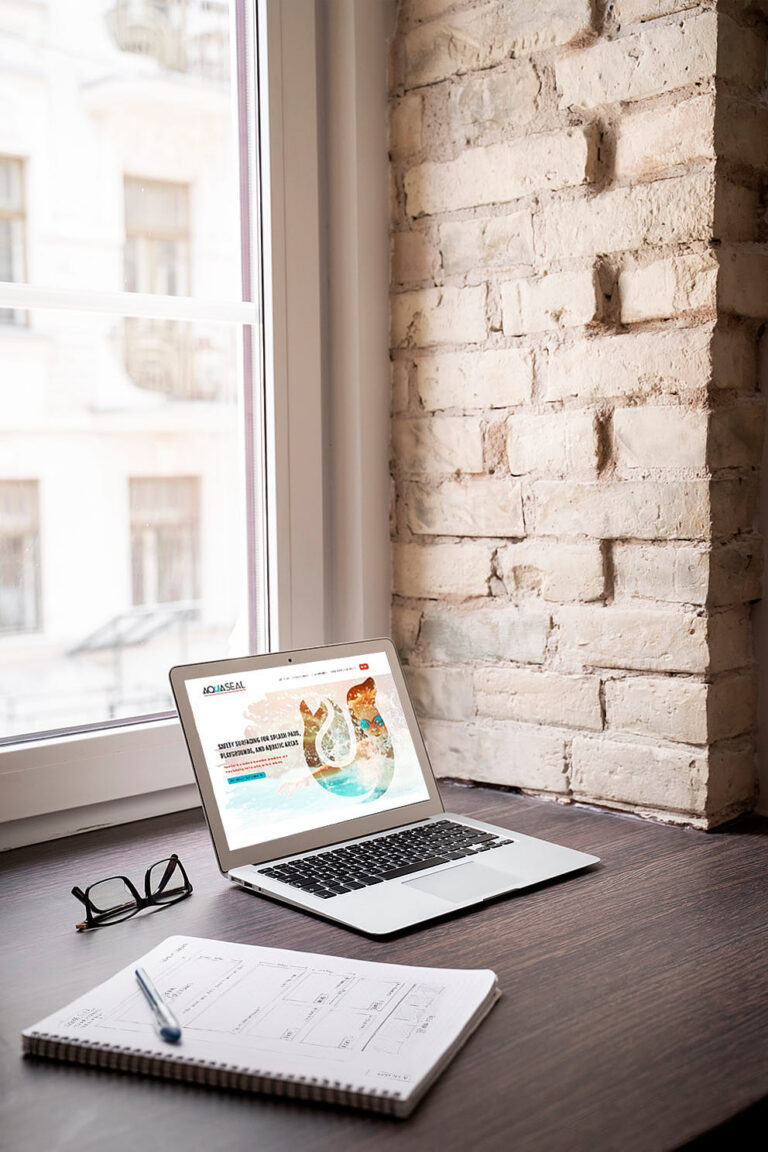 Laptop on a desk by a window displaying a website design, with a notebook, pen, and glasses nearby.