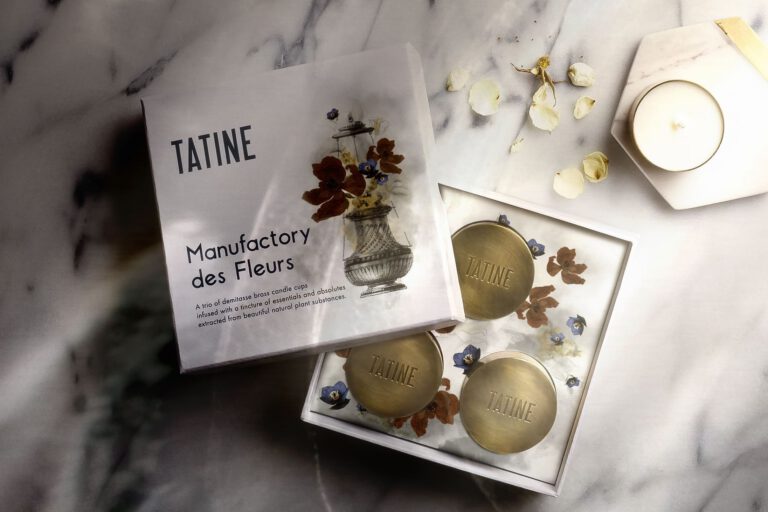 Tatine ‘Manufactory des Fleurs’ gift set box shown open on a marble surface, revealing three engraved brass candle cups on a floral-illustrated insert, with scattered white petals and a small white candle nearby. Package designs by Rebecca Snyder, an award winning graphic designer, and owner of A LA MODE designs, a graphic studio located in Canton, Ohio.