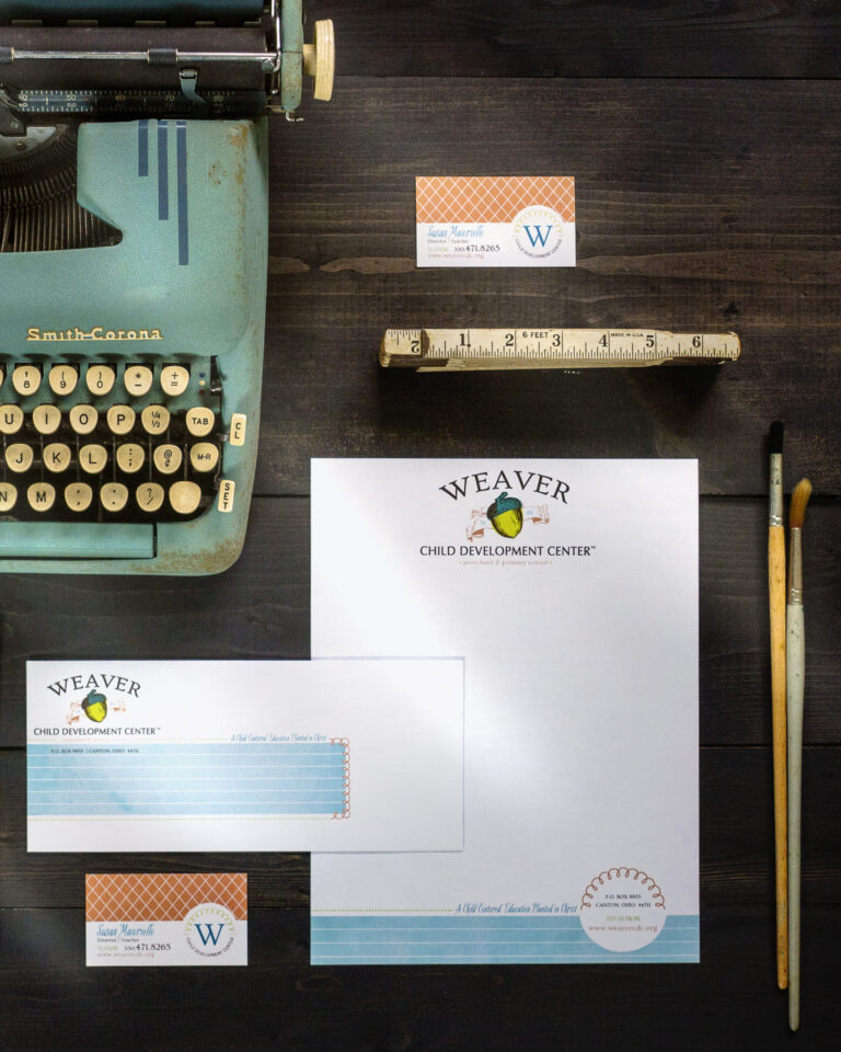 Weaver Child Development Center stationery set—letterhead, envelopes, and business cards—arranged on a dark wood surface beside a vintage typewriter, ruler, and paintbrushes. Brand identity by A LA MODE designs, Rebecca Snyder, lead graphic designer, owner.