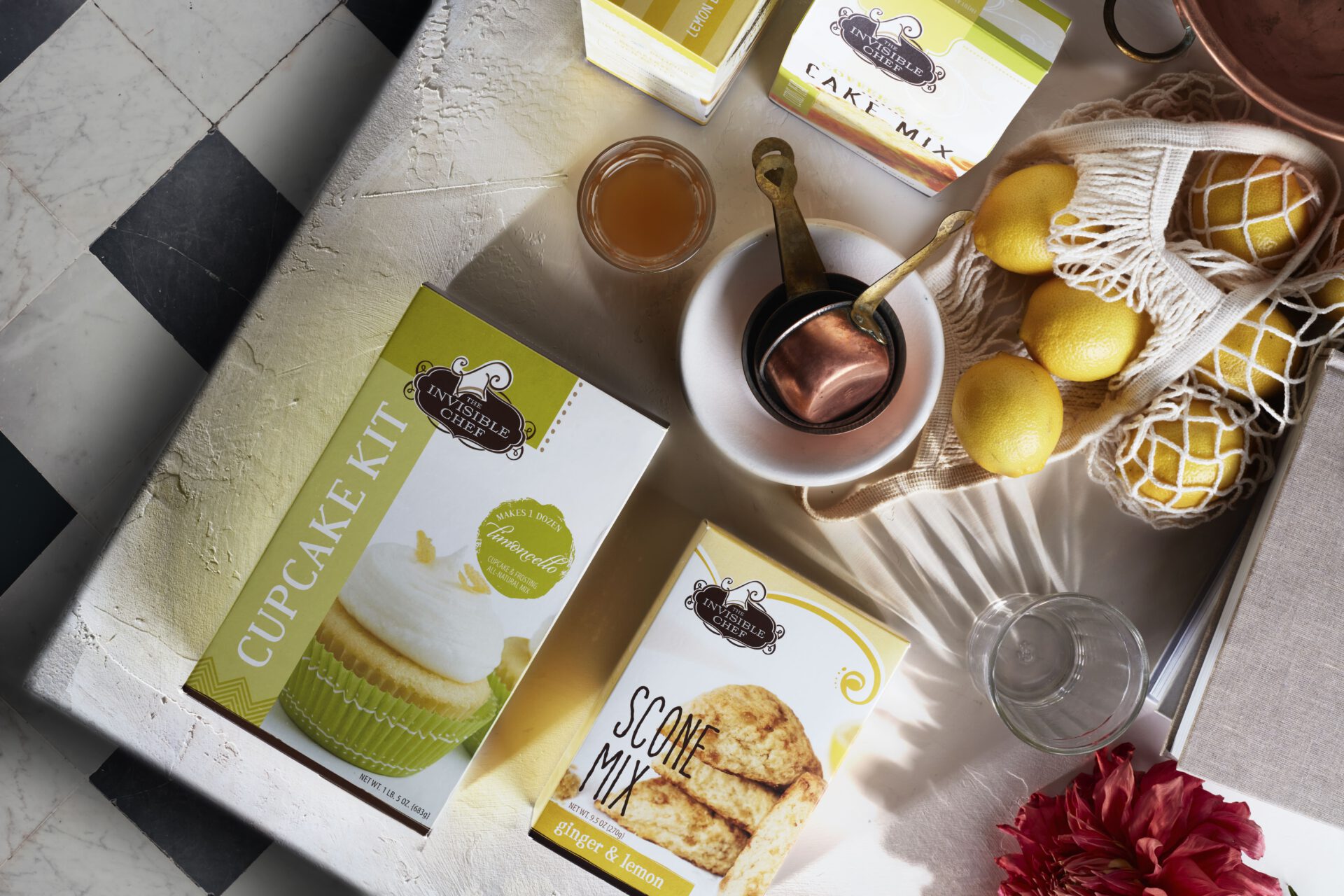 Flat-lay of The Invisible Chef baking mix packaging (“Cupcake Kit” and “Scone Mix”) styled with fresh lemons and kitchen tools. Packaging design by Rebecca Snyder at A LA MODE designs.