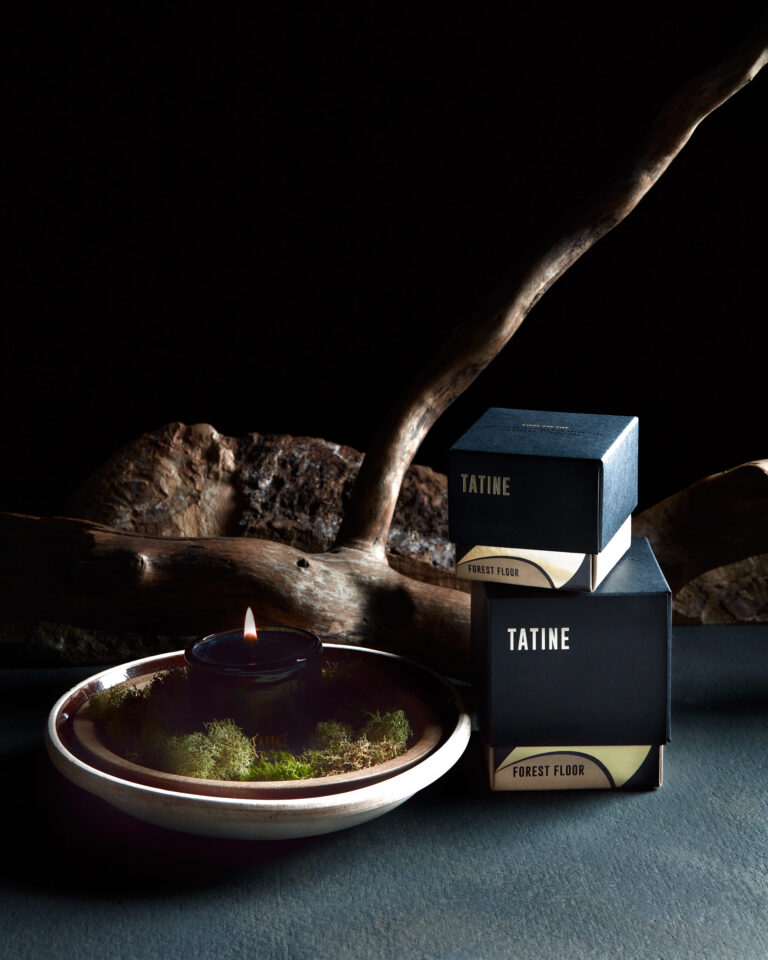 Moody still life of a lit Tatine candle in a shallow bowl with moss, beside two stacked Tatine ‘Forest Floor’ boxes, set against driftwood and a dark background. Package design services by A LA MODE designs, an award-winning graphic design studio, located in Canton, Ohio. Rebecca Snyder, leader designer, and owner.
