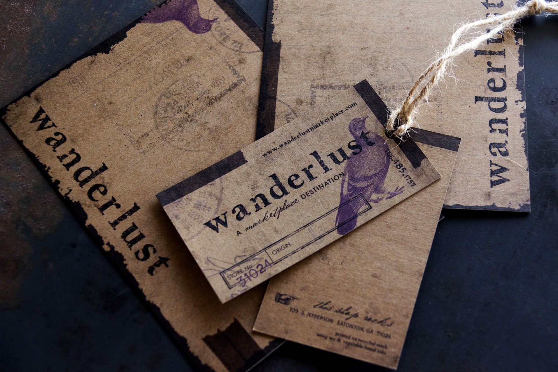 Close-up of rustic kraft-paper hang tags and a matching Wanderlust business card, featuring stamped typography, a purple bird illustration, and twine tie. Print and brand identity by Rebecca Snyder, an award-winning graphic artist for A LA MODE designs.