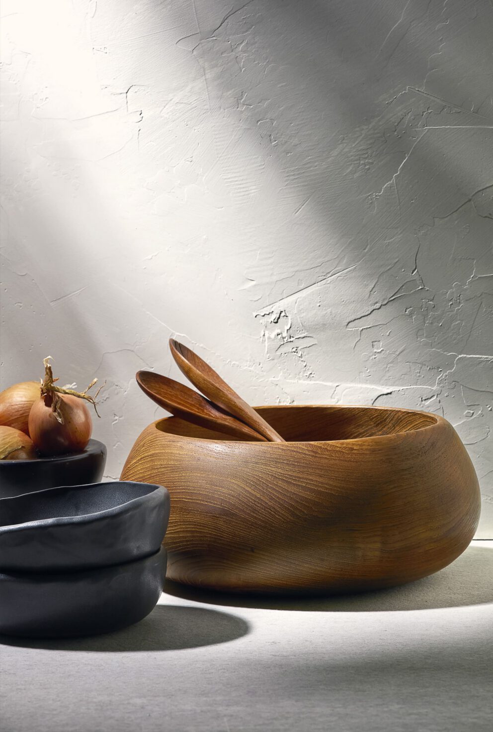 Still-life of a wooden bowl with wooden spoons beside dark ceramic bowls and an onion, lit by sunlight against a textured wall. Photography styling and services by A LA MODE designs.