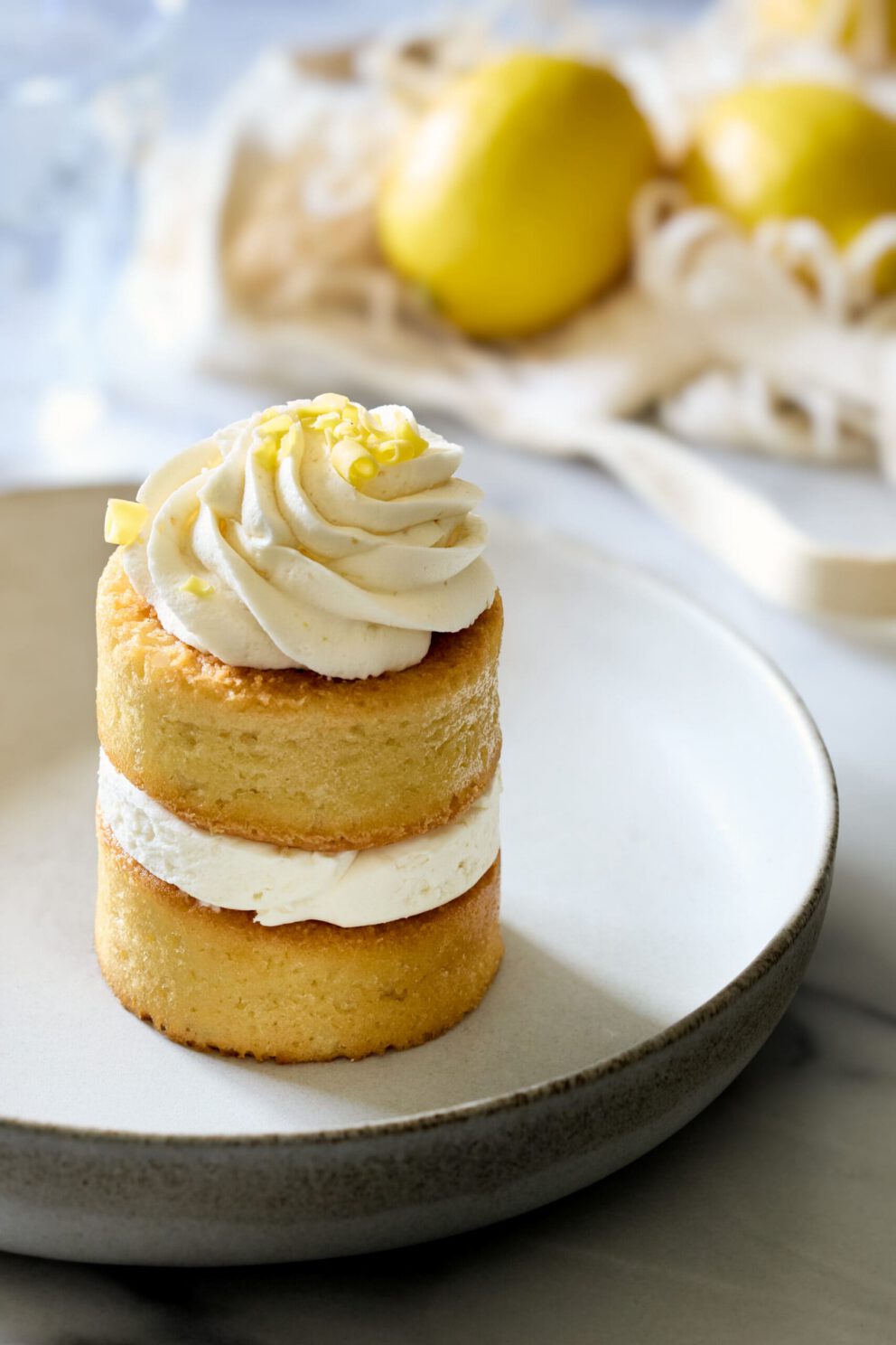 Mini layered lemon cake with whipped frosting on a plate, with lemons in the background. Photography and styling services provided by A LA MODE designs, Matt & Rebecca Snyder.